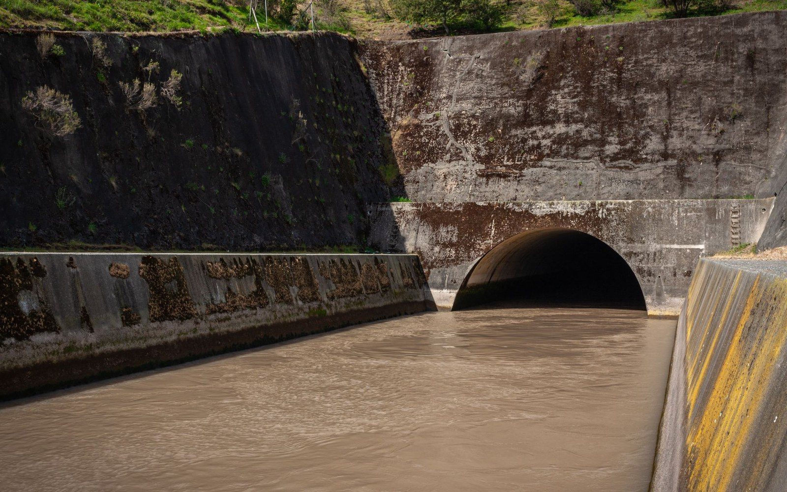 Muddy water flows through a concrete spillway channel into an arched tunnel outlet