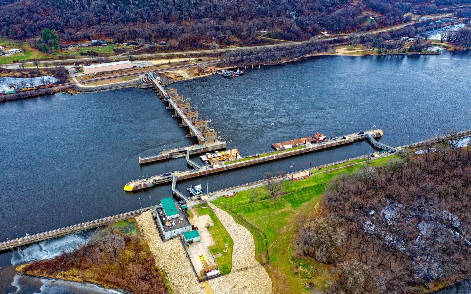 Aerial view of a river dam and lock system with a barge alongside wooded banks