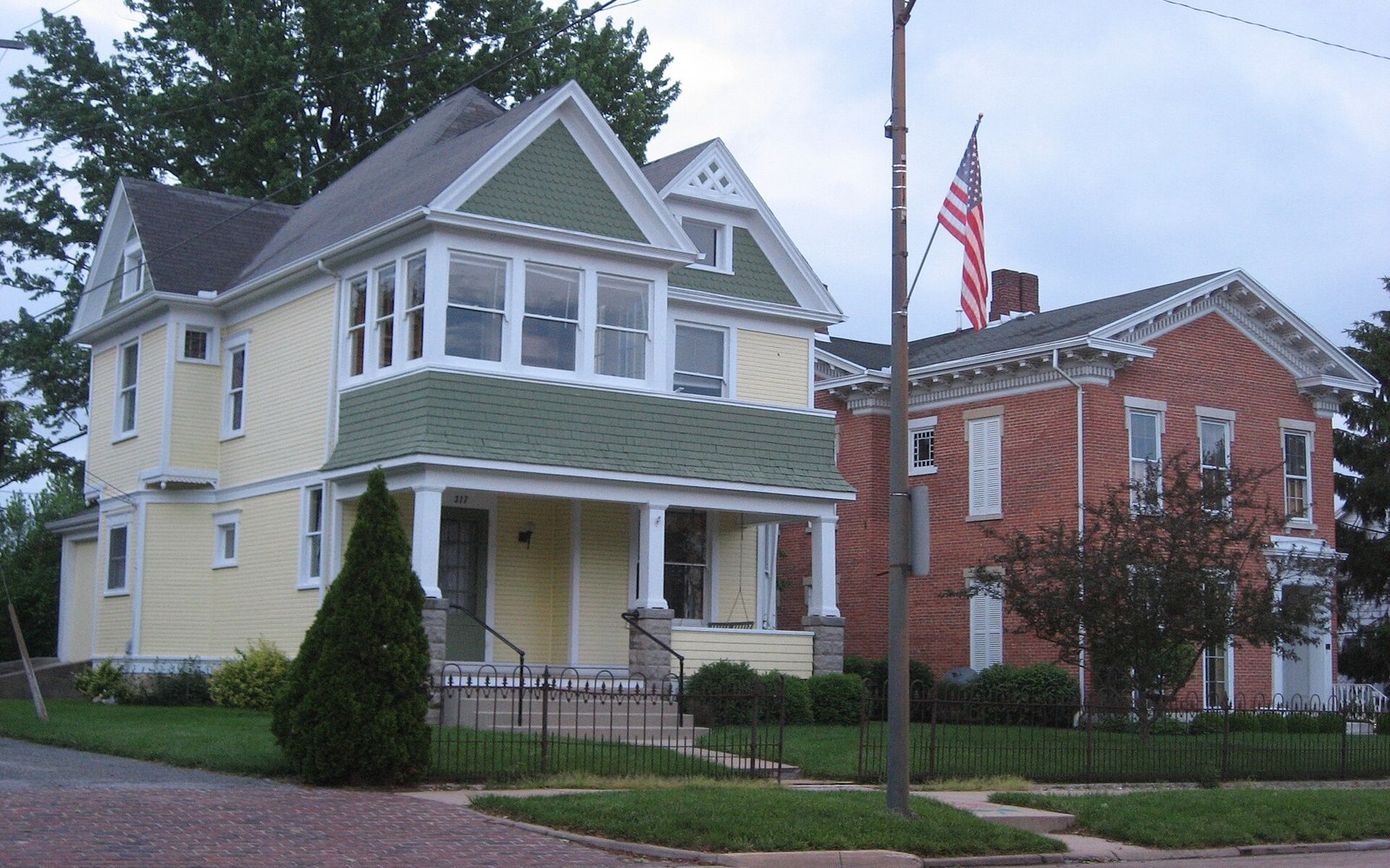 Victorian yellow house with a bay window stands beside a brick house flying an American flag