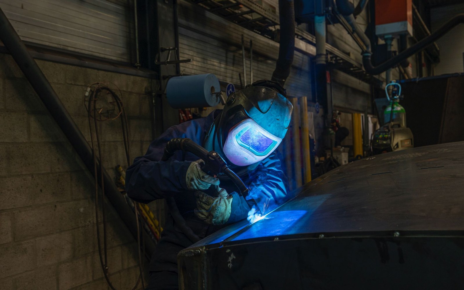 Hobart Institute of Welding Technology training bay: adult student in helmet and leathers, live welding arc, instructor standing behind
